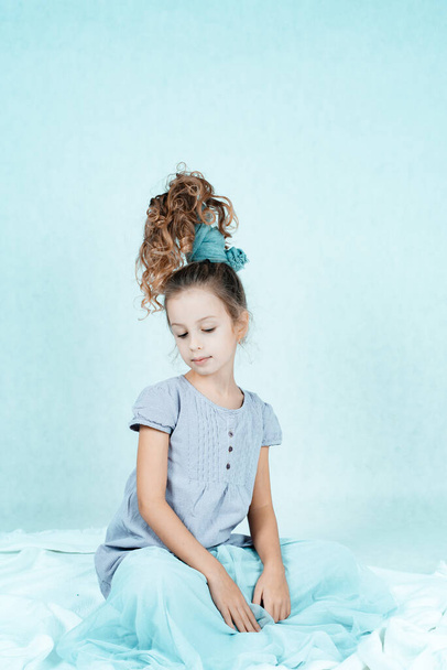 A cute little girl with long dark hair gathered in a ponytail in a gray dress poses on a white background in the studio. The child is isolated on a white background. A child with curly hair. A girl with an unusual hairstyle. - Foto, Bild