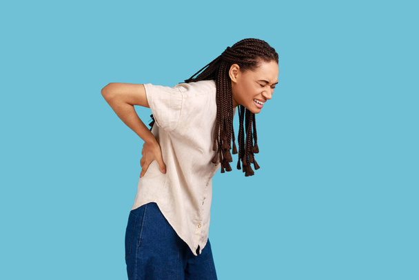 Woman with black dreadlocks has backache, suffers from painful muscle injury, smirks face from unpleasant feelings, wearing white shirt. Indoor studio shot isolated on blue background. - Фото, зображення