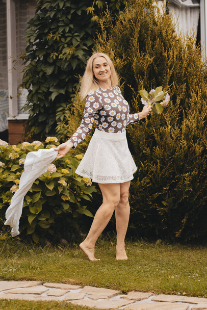 Happy woman with blond hair in a white skirt and a summer sweater walks barefoot in the park. Girl holding flowers outdoors. Happy girl enjoying the warm summer weather. The girl runs on the lawn. - Photo, Image