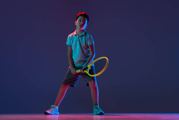 Full-length portrait of boy, tennis player posing isolated over blue background in neon lights - Foto, immagini