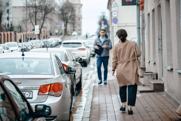 Minsk, Belarus-April 08, 2021: A young man walks down the street along houses and cars with documents in his hands. An attractive girl in a coat walks around the city. - Zdjęcie, obraz