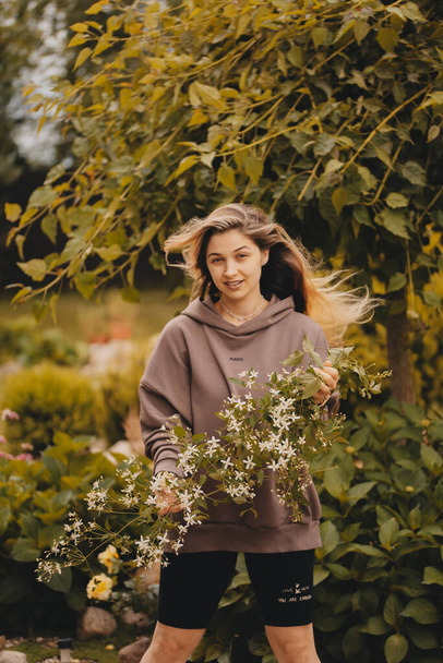 A beautiful young girl with long hair in a brown sweater posing in nature with flowers in her hands. Happy girl enjoying the warm summer weather. Long hair fluttered in the wind. - Foto, Imagem