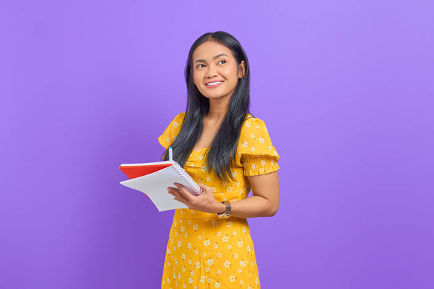 Sorrindo jovem mulher asiática segurando caneta e caderno, olhando para cima isolado no fundo roxo - Foto, Imagem