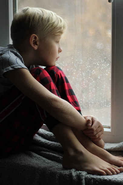 Bir çocuk pencereden dışarı bakıyor. Klasik renk. turuncu lastik çizmeler pencerenin yanında duruyor - Fotoğraf, Görsel