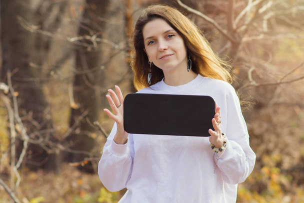Mockup with a beautiful young brunette girl in a white sweater holding a black sign with space for text. A woman in the park on a warm autumn day with a board in her hands. Copyspace - Φωτογραφία, εικόνα