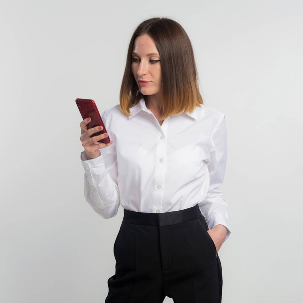 Portrait of pretty, stylish woman in denim outfit holding smart phone in hands, checking email, searching, using internet, chatting with friends standing on grey background, copy space - Zdjęcie, obraz