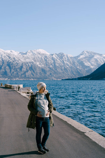 Raskaana oleva nainen takissa seisoo asfaltin penkereellä ja katsoo merta - Valokuva, kuva