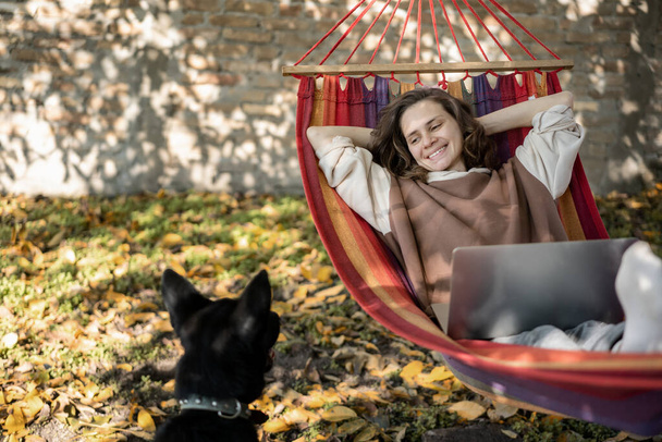 Young happy woman freelancer with laptop and dog in hammock at backyard of country house on autumn day. - Foto, Imagen