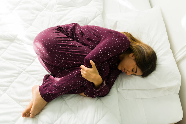 Νιώθω τόσο άσχημα. Νεαρή γυναίκα με κράμπες που αγκαλιάζει τα γόνατά της στο κρεβάτι και υποφέρει από στομαχόπονο - Φωτογραφία, εικόνα