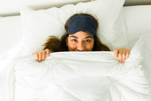 I love my bed. Happy young woman smiling under the covers in bed with a sleep mask  - Photo, Image