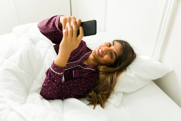 豪華なヒスパニック女性のテキストメッセージと彼女のスマートフォンでソーシャルメディアを使用しながら、白い快適なベッドの上に休んで - 写真・画像