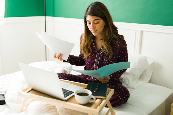 ベッドの中の魅力的な女性は、自宅からリモートで作業しながら、ノートパソコンを使用して作業用紙を保持  - 写真・画像