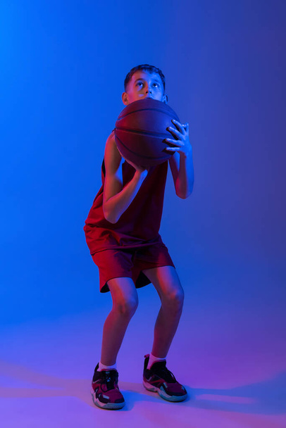 Full-length portrait of basketball player training, throwing ball into basket isolated over gradient blue purple background in neon lights - Photo, Image