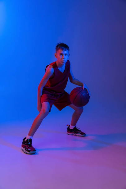 Full-length portrait of boy, basketball player in motion, training isolated over gradient blue purple background in neon lights - 写真・画像