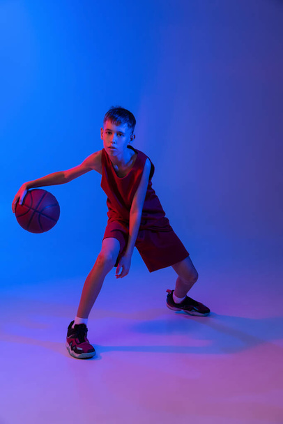 Portrét profesionálního teen basketbalista v pohybu izolované přes gradient modré pozadí v neonovém světle - Fotografie, Obrázek