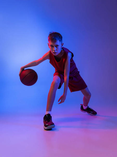 Full-length portrait of basketball player training, dribbling isolated over gradient blue background in neon light - Foto, Bild