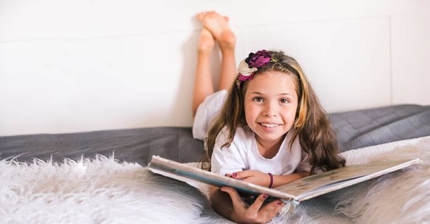 Schoolgirl reading a book in the room. Girl 8 or 10 years old with a book in the room on the bed. A child in a bright interior to study at home - Φωτογραφία, εικόνα