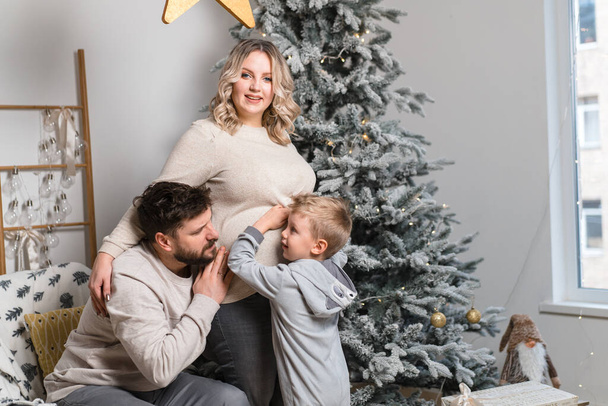 Christmas Family Happiness Portrait of dad, pregnant mom and little son  sitting armchair at home near Christmas tree hug smile European young adult family holiday morning - Фото, изображение