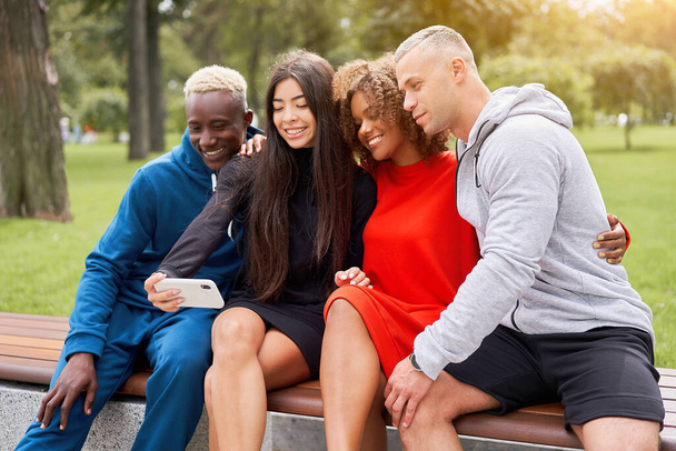 Young people of different nationalities take selfies using smartphones, sitting on a bench in the park in summer. Athletic build. Sports uniform. Young happy students. - 写真・画像