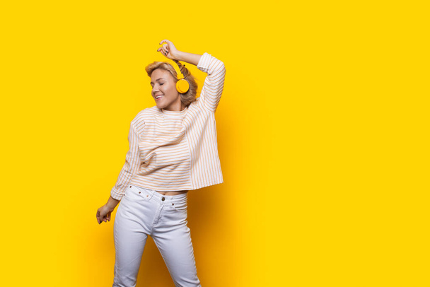 Mujer caucásica alegre levanta la mano en movimiento de baile mientras escucha música en los auriculares. Aislado sobre el patio amarillo con espacio de copia - Foto, imagen