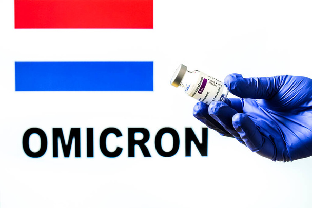 Madrid, Spanyolország - 2021. november 29.: Kézben tartó injekciós üveg Astrazeneca covid vakcinával. Holland zászló és Omicron kovid változat szó írva a háttérben - Fotó, kép