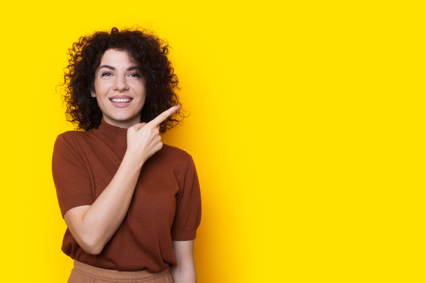 Front view of a happy woman standing isolated over yellow background looking at camera and pointing with index finger at emty space. Copyspace. Curly hair - Foto, afbeelding
