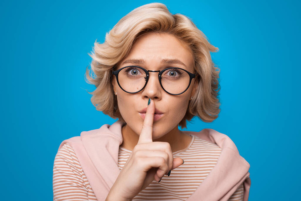 Mujer rubia sorprendida hace gesto de silencio presiona el dedo índice sobre los labios aislados sobre fondo azul. Señal Shhh. - Foto, imagen