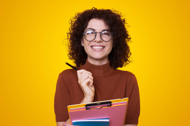 Porträt einer Frau, die Werbebücher in der Hand hält, zahm lächelnd in die Kamera blickt, isoliert auf gelbem Hintergrund. - Foto, Bild