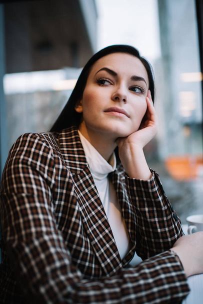Portrait of young pensive female in checkered jacket with dark hair looking away while leaning on hand sitting at table - Фото, зображення