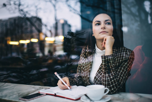 Through window of thoughtful female remote worker looking away while sitting at table leaning on hand and writing notes during work - Фото, изображение