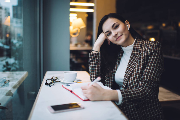 Smiling young dark haired businesswoman in formal clothes and earphones sitting at table in with coffee and smartphone while writing notes in notebook - Foto, Bild
