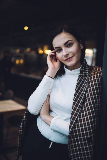 Улыбающаяся молодая женщина в модном пальто и белой водолазке, опираясь на стену, стоя в кафетерии, трогая волосы и глядя в камеру - Фото, изображение