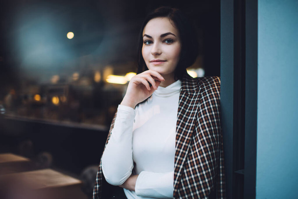 Smiling young woman wearing white turtleneck and stylish coat leaning on blue wall in cafeteria while touching chin and looking at camera - Foto, afbeelding