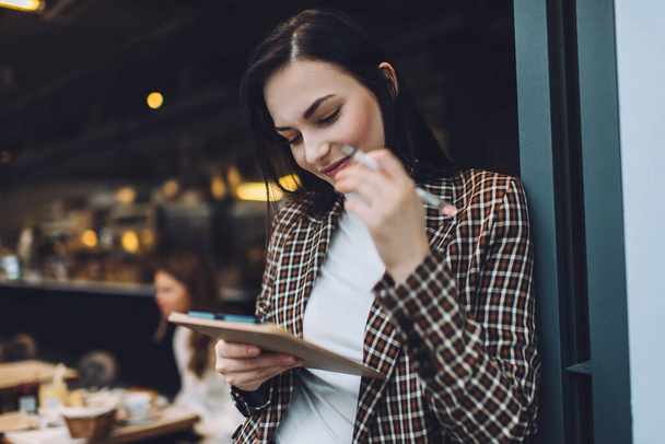 Confident young woman in elegant outfit standing with clipboard and making notes with pen while leaning on cafe wall in day - Фото, изображение