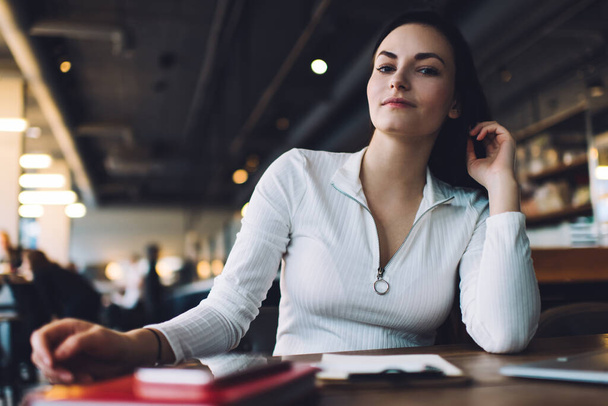 Calm female freelancer in casual clothes looking at camera while sitting at table in cafeteria with clipboard working on project remotely - Foto, Bild