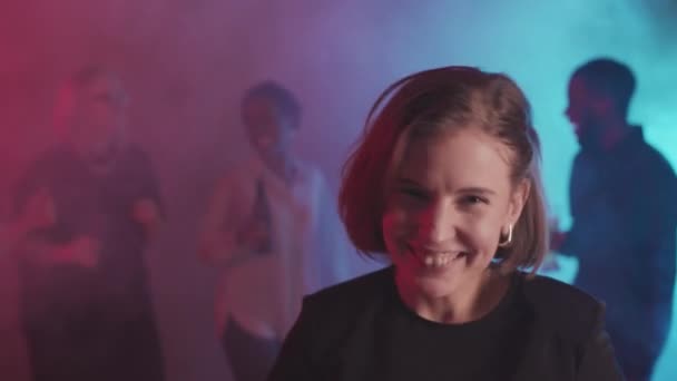 Medium close-up met slow mo van jonge vrolijke vrouw met een glas rode wijn energiek dansen in rokerige kamer met paarse verlichting tijdens het feest. Mensen clubben op de achtergrond - Video