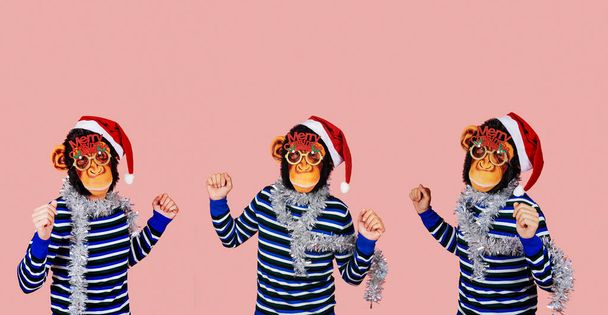 tres hombres bailando con máscaras de mono, sombreros de santa, anteojos con la frase feliz Navidad, y tiras de oropel de plata alrededor de sus cuellos, sobre un fondo rosa con un poco de espacio en blanco en la parte superior - Foto, imagen