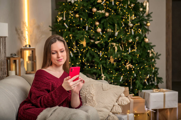 A young beautiful girl sits on a cozy sofa and looks surprisingly into a smartphone, against the background of a Christmas decorated Christmas tree - Zdjęcie, obraz