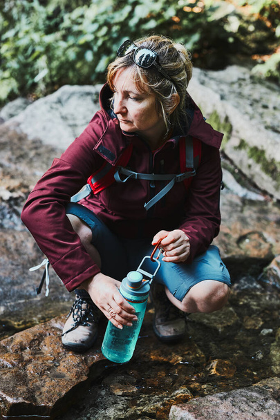 Woman taking pure water to bottle from mountain stream during trekking in mountains. Hiker crouching on rocks, filling bottle up with cold mountain water. Enjoying the outdoors in the summer trip vacation - Fotoğraf, Görsel