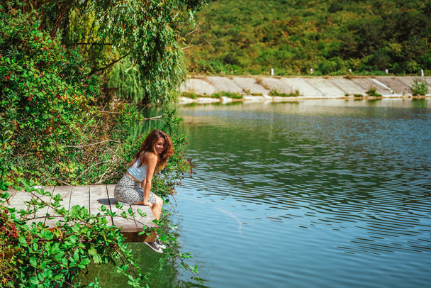  スカートに身を包んだ魅力的な若い女性が、森の中の湖を背景に木の橋の上に腰を下ろしている。旅行、自由、アクティブなライフスタイルの概念. - 写真・画像