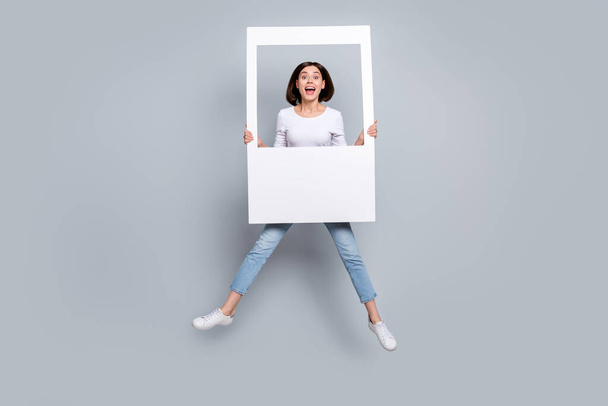 Photo of sweet impressed young woman wear white outfit jumping high holding white photo frame smiling isolated grey color background - Foto, Bild