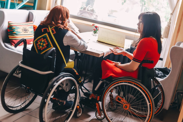 Δύο κορίτσια σε αναπηρικά καροτσάκια σε ένα ζεστό καφέ περιμένουν για μια παραγγελία. Προσβάσιμοι κοινοί χώροι για άτομα με αναπηρία. Προσιτός τουρισμός για άτομα με ειδικές ανάγκες λόγω σωματικής αναπηρίας - Φωτογραφία, εικόνα