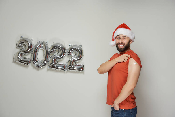 Ein glatzköpfiger Mann mit Bart und Weihnachtsmütze lächelt und zeigt Schulter mit Pflaster nach Impfung gegen Coronavirus (Covid-19) neben vereitelten silbernen Luftballons in Form von 2022.  - Foto, Bild