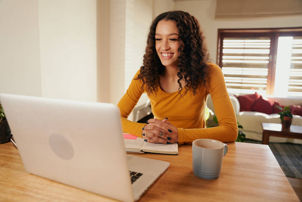 Mulher multicultural sorrindo enquanto em reunião online. Jovem profissional remoto trabalhando com laptop no apartamento moderno - Foto, Imagem