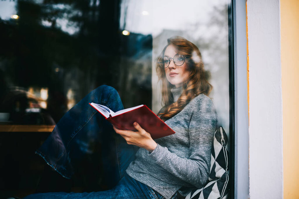Through window of thoughtful female with eyeglasses and wavy hair looking away sitting on windowsill in comfortable pose and reading book having free time - Фото, зображення