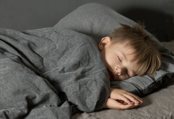 a 5-year-old preschooler boy sleeps in bed on a pillow under a blanket. Grey dark background - Foto, afbeelding