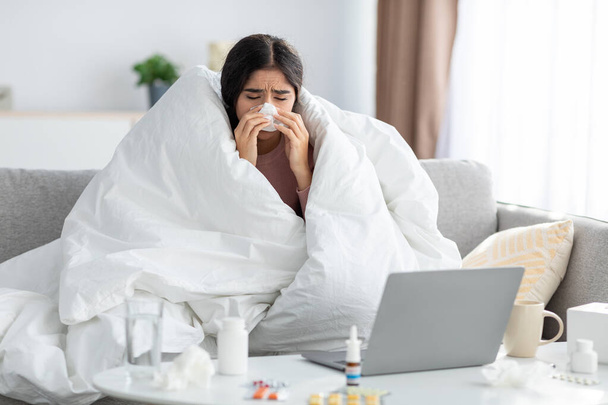 Frustration jeune dame indienne enveloppée dans la couverture souffle nez dans la serviette sur le canapé avec ordinateur portable et la médecine - Photo, image