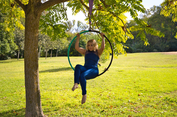 Blonde woman and young gymnast acrobat athlete performing aerial exercise on air ring outdoors in park. Lithe woman in blue costume performs poses of circus performers dancing with hips. - 写真・画像