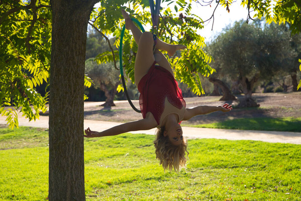 Blonde woman and young gymnast acrobat athlete performing aerial exercise on air ring outdoors in park. Flexible woman in red costume performs poses of circus performers dancing with hips. - Photo, Image