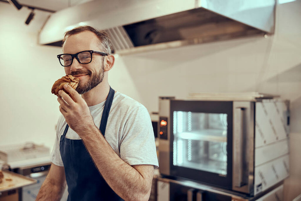 Bearded man in apron smelling pastry in cafe - Zdjęcie, obraz